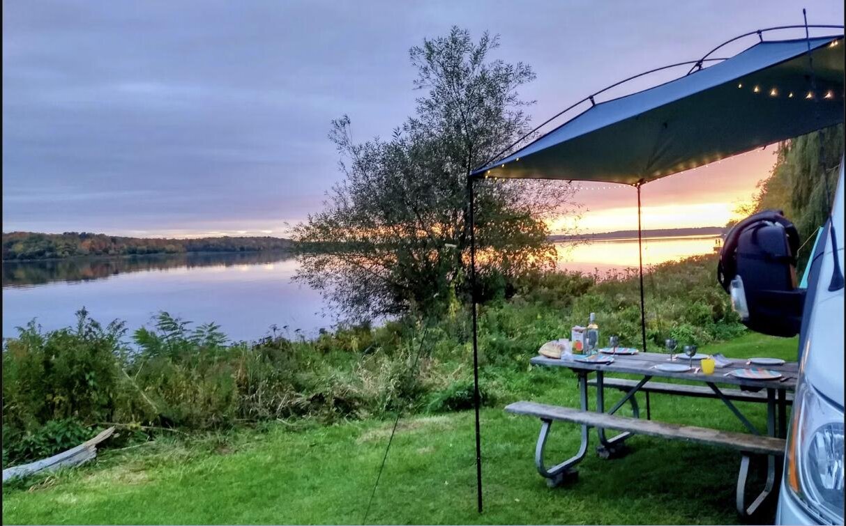 Campervan parked at a lakeside campsite at golden hour, picnic table and string lights set up alongside