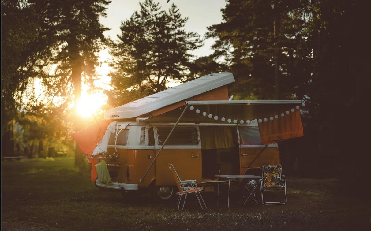 Classic yellow campervan with a pop-top roof parked in a forest clearing at sunset, camp chairs set up alongside